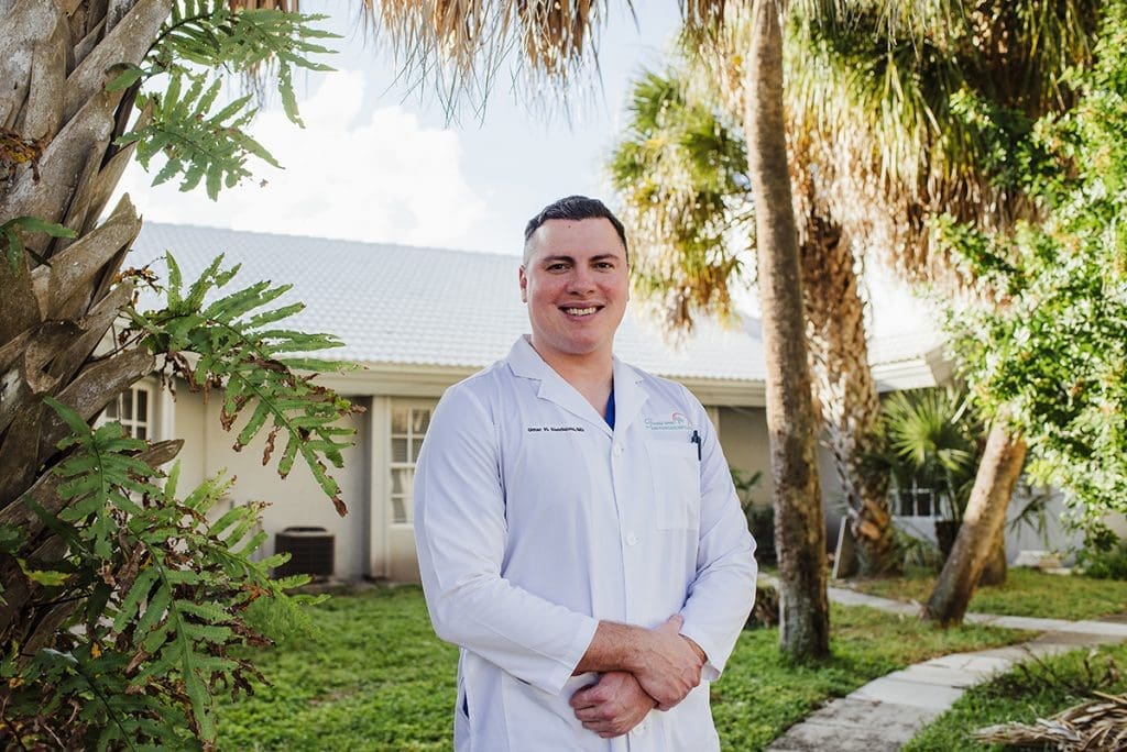 A man in a white coat, specializing in non-surgical pain treatment, standing in front of a palm tree.