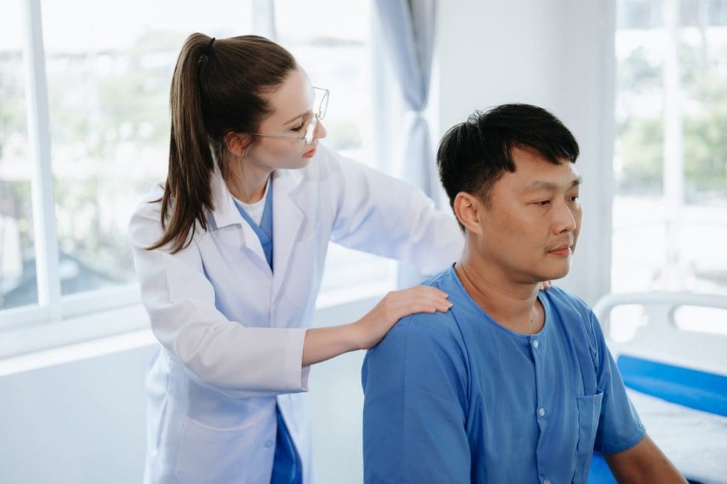 Female physiotherapist in lab coat examining the back of a male patient seated in a clinic.