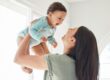 A woman smiles and lifts a happy baby wearing a light blue onesie in a bright room.