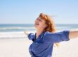 A woman with red hair smiles with outstretched arms on a sandy beach with the ocean and blue sky in the background. Chronic Joint Pain